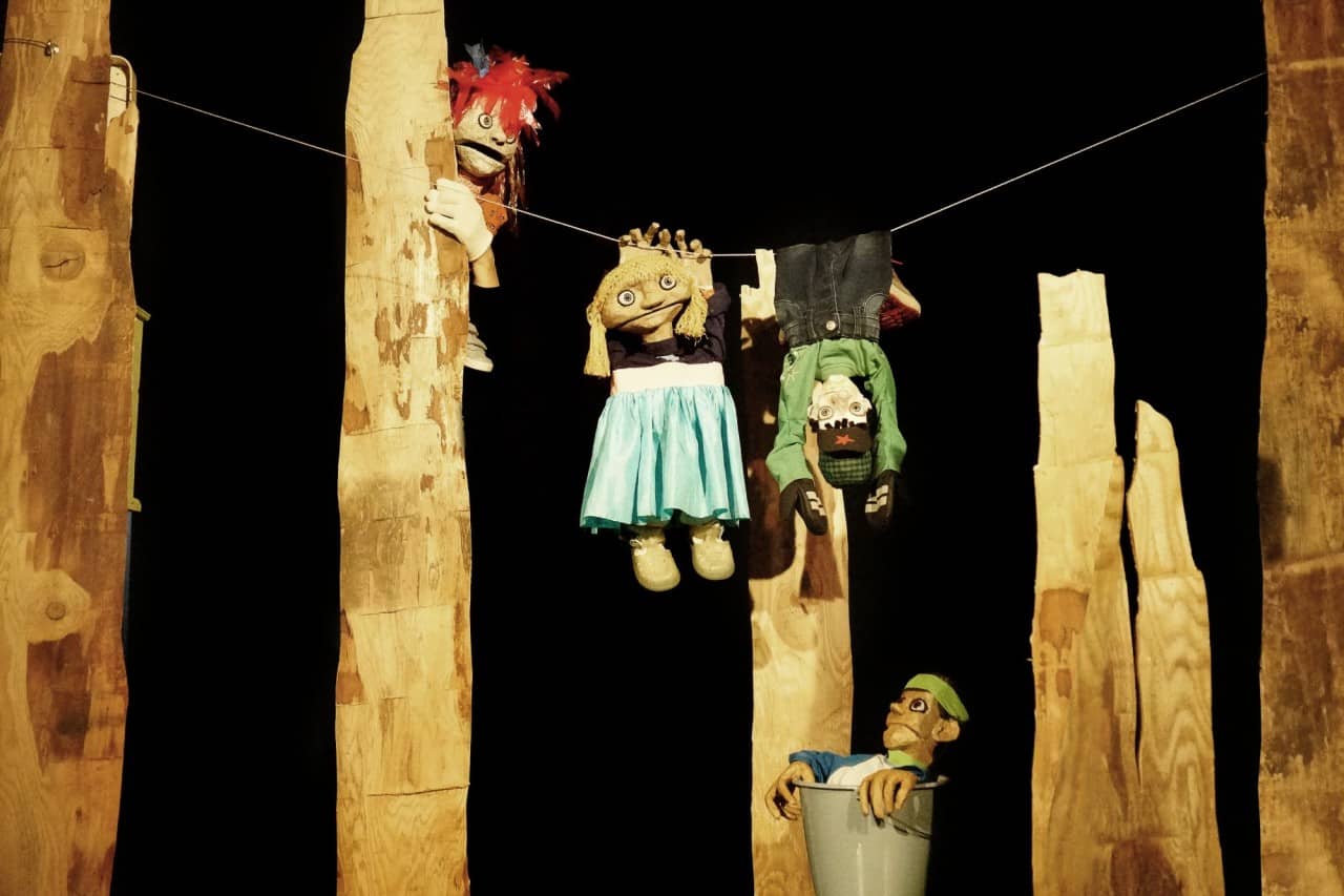 Vier Puppenspielfiguren hängen an einer Schnur, sitzen in einem Eimer oder schauen hinter einem Holzstück hervor.