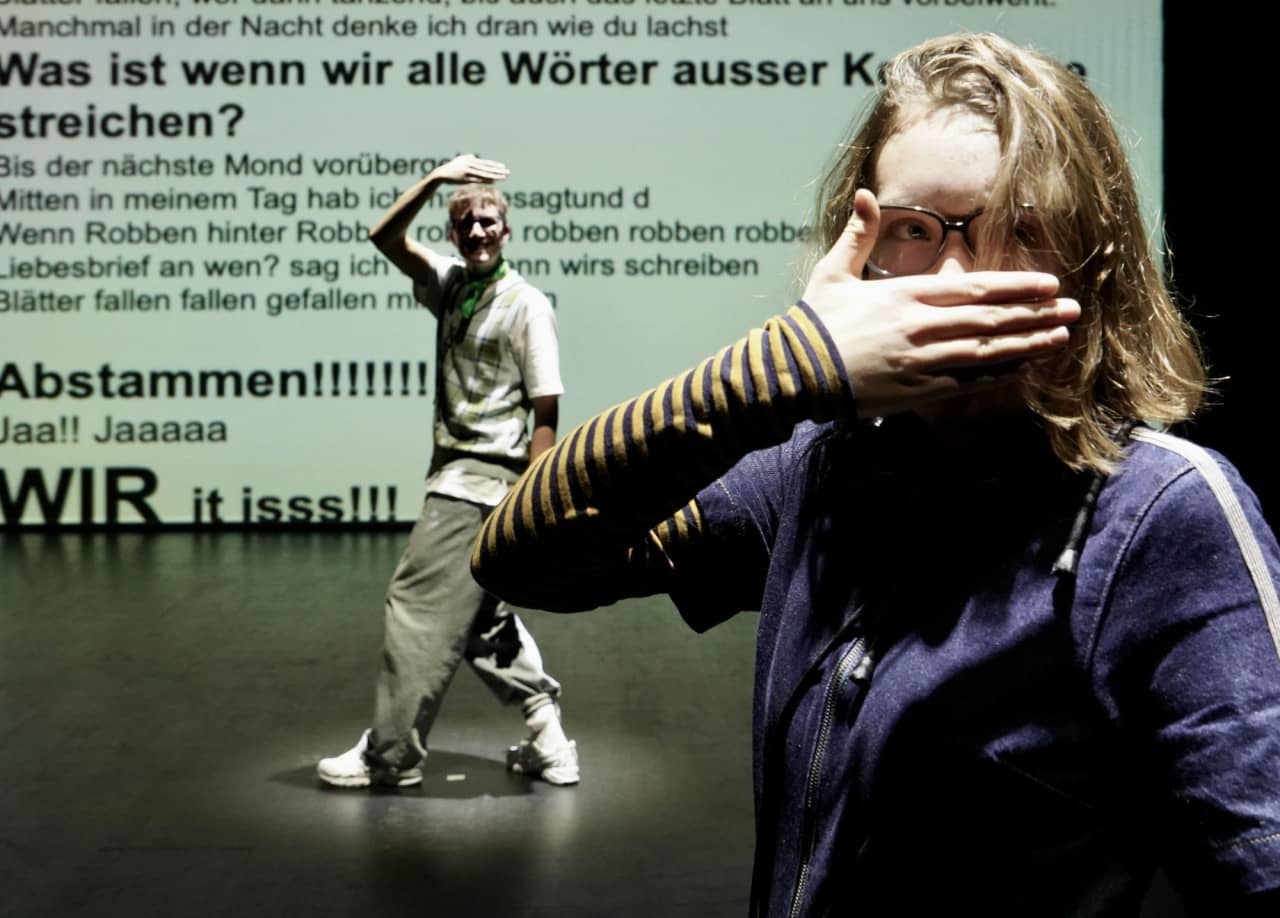 Zwei Jugendliche mit Hand vor dem Mund und im Hintergrund ein Text an der Wand