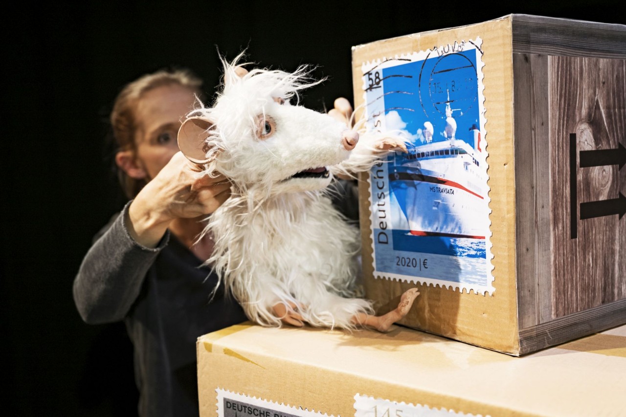 Eine weisse Tierpuppe befindet sich neben zwei Paketkisten mit aufgedruckten Briefmarken und Pfeilsymbolen; im Hintergrund ist eine Person zu sehen, die die Tierpuppe führt.