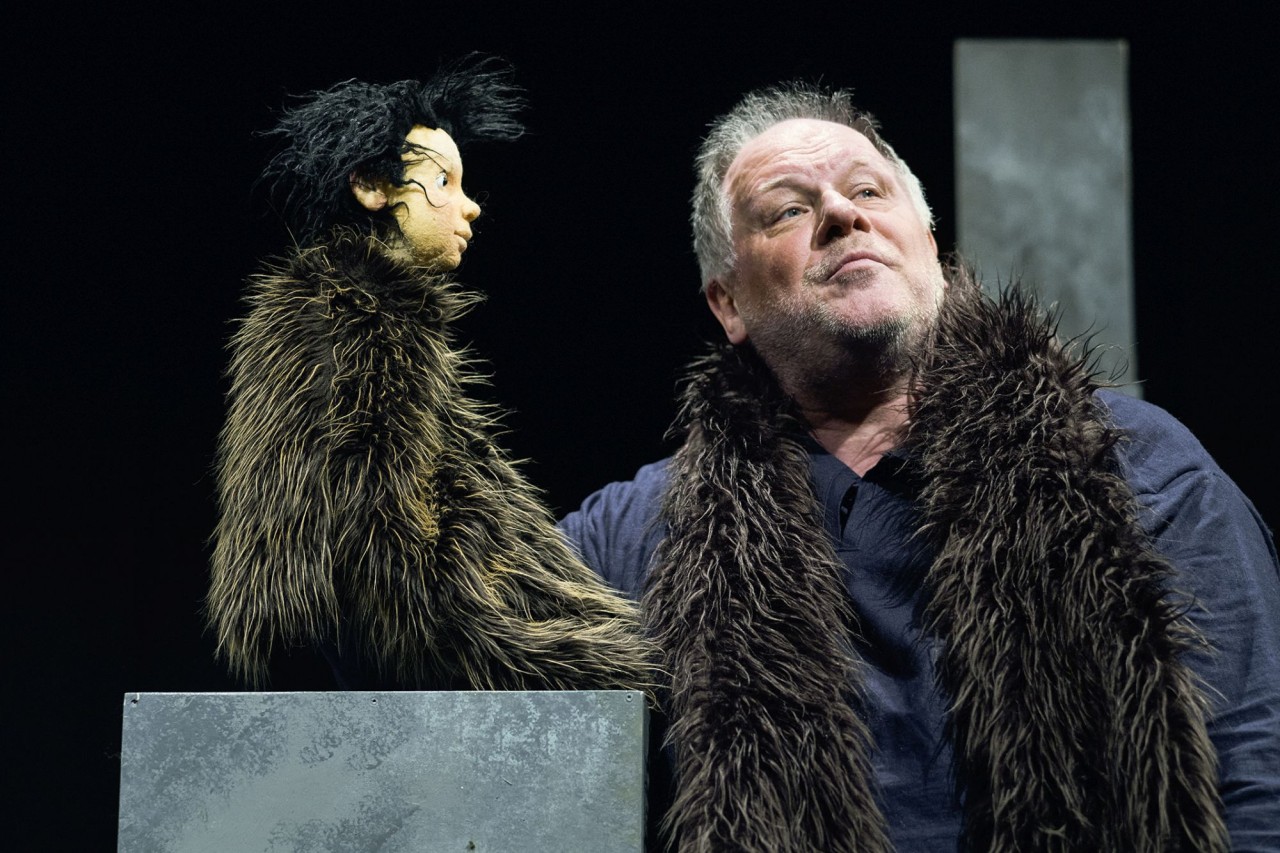 Puppenspieler mit einer grossen dunklen, zotteligen Tierfigur auf einer Theaterbühne vor schwarzem Hintergrund.