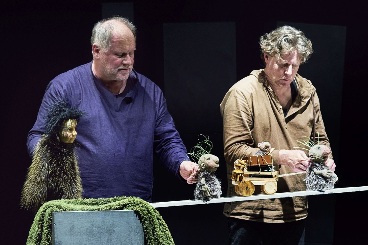 Zwei Puppenspieler auf einer dunklen Bühne führen kleine mausähnliche Figuren und einen kleinen Holzwagen in einer Theaterszene.
