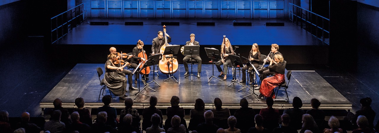 Ein Kammermusikorchester bestehend aus neun Musikerinnen und Musikern auf einer Bühne.