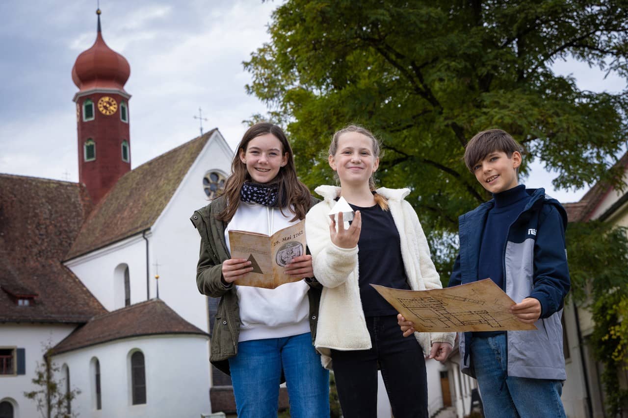 Drei Kinder stehten vor dem Kloster Wettingen, in der Hand halten sie Utensilien für die Rätseltour: Pyramide, Karte und Buch