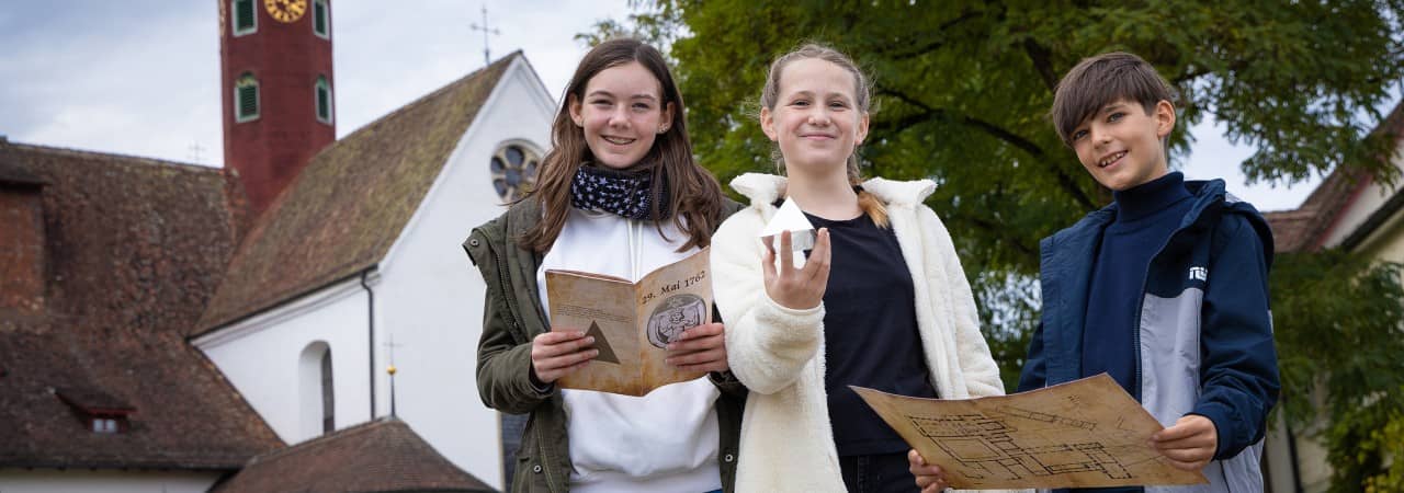 Drei Kinder stehten vor dem Kloster Wettingen, in der Hand halten sie Utensilien für die Rätseltour: Pyramide, Karte und Buch