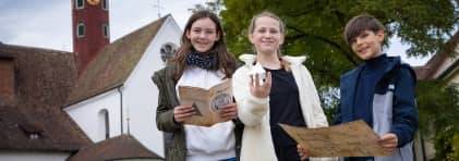 Drei Kinder stehten vor dem Kloster Wettingen, in der Hand halten sie Utensilien für die Rätseltour: Pyramide, Karte und Buch