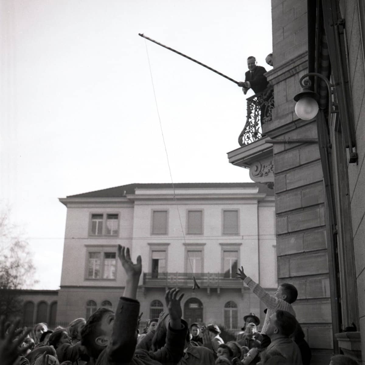 Ein Mann steht auf einem Balkon mit einer Angelrute, unter ihm steht eine Memge von Kindern, die etwas aufzufangen versuchen. 