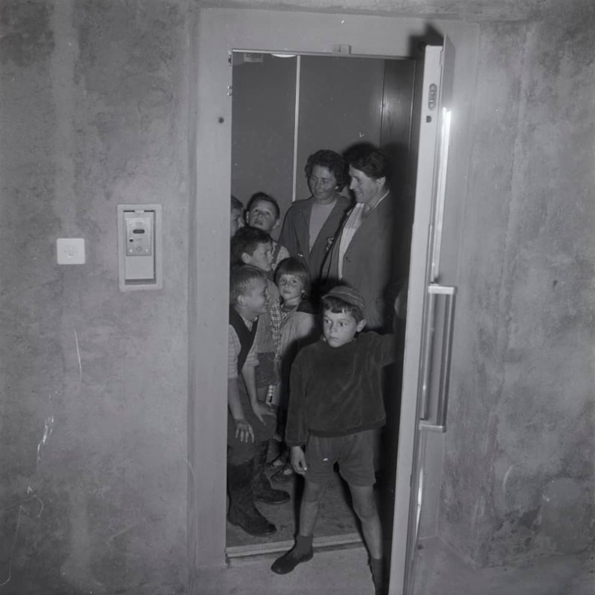Erwachsenen und Kinder in einem Lift, ein Knabe im Vordergrund schliesst die Lifttür von innen. Aarburg, August 1960.