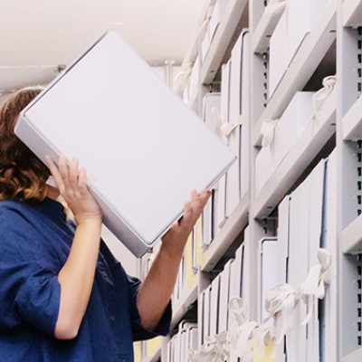 Zwei Archivregale, die in der Mitte des Bildes beinahe in der Flucht zusammenlaufen. Zwischen den Regalen auf einem roten Tritthocker eine Frau mit dunklen, schulterlangen Haaren, die eine Archivschachtel auf der Höhe ihres Kopfes hält. Sie ist im Begriff, diese wieder zu verräumen.