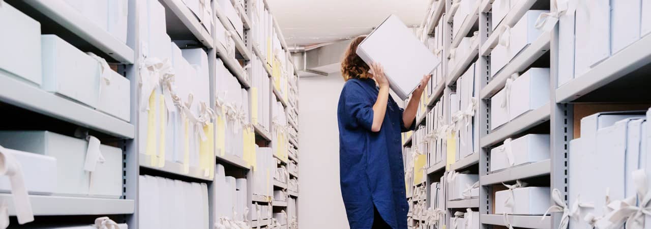 Zwei Archivregale, die in der Mitte des Bildes beinahe in der Flucht zusammenlaufen. Zwischen den Regalen auf einem roten Tritthocker eine Frau mit dunklen, schulterlangen Haaren, die eine Archivschachtel auf der Höhe ihres Kopfes hält. Sie ist im Begriff, diese wieder zu verräumen.