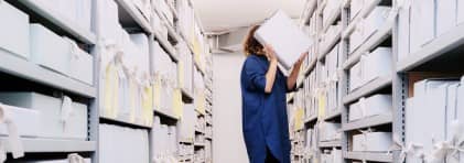 Zwei Archivregale, die in der Mitte des Bildes beinahe in der Flucht zusammenlaufen. Zwischen den Regalen auf einem roten Tritthocker eine Frau mit dunklen, schulterlangen Haaren, die eine Archivschachtel auf der Höhe ihres Kopfes hält. Sie ist im Begriff, diese wieder zu verräumen.