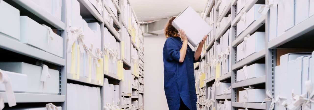 Das Foto zeigt eine Person, die auf einem Schemel steht und im Staatsarchiv eine Archivschachtel aus dem Regal nimmt.
