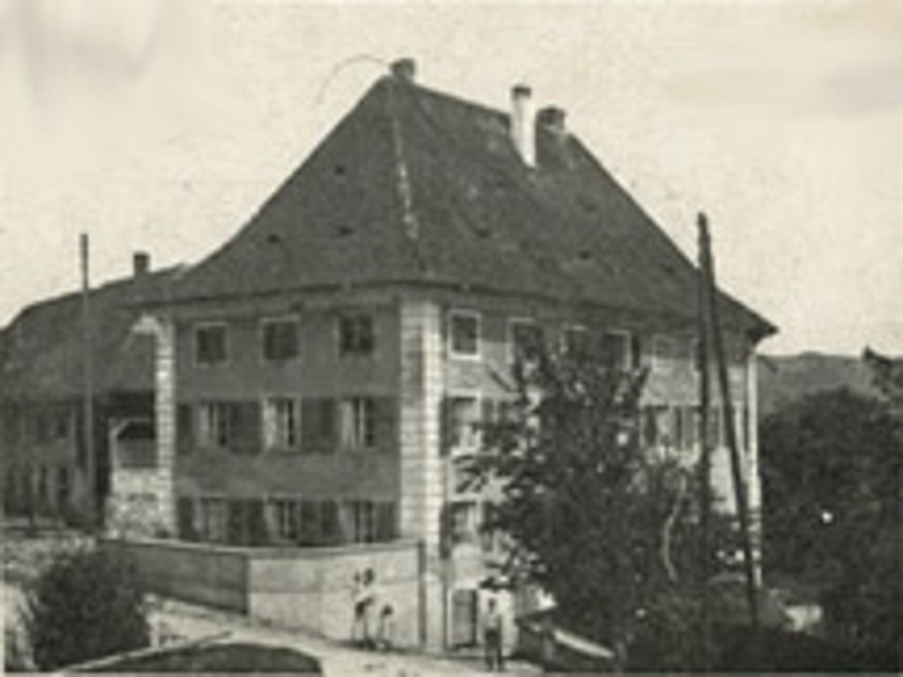 Schwarz-weiss Fotografie von Aussenansicht eines mehrstöckigen Schulhauses mit einem Baum und Personen im Vordergrund.