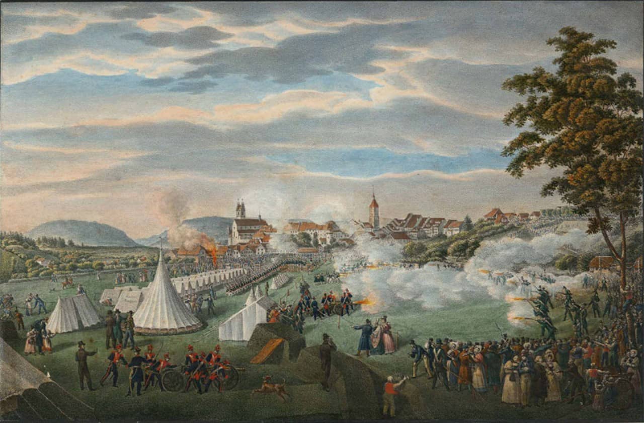 Kolorierte Zeichnung auf Papier. Im Vordergrund ist ein Zeltlager mit vielen Personen die bei Schiessübungen von Kadetten zusehen. Rechterhand ist ein Baum zu sehen und im Hintergrund die Stadt Aarau und Hügellandschaft. Knapp die Hälfte des Bildes nimmt ein grau-blauer Wolkenverhangener Himmel ein.