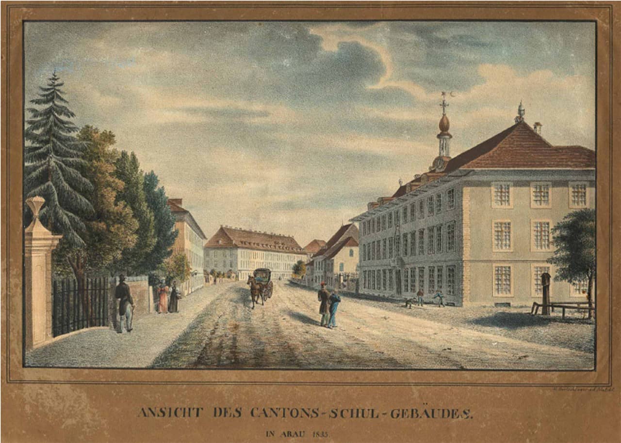 Kolorierte Zeichnung auf Papier mit braunem Rahmen. Untenstehend gedruckter Titel "Ansicht des Cantons-Schul-Gebäudes in Aarau 1835". Rechts einer grossen Strasse ist das Kantonsschulgebäude zu sehen. Auf der Strasse und dem Trottoir sind verschiedene Menschen und eine Pferdekutsche abgebildet. Links wird die Strasse von Bäumen hinter einem Gartenzaun gesäumt. An der Strasse stehen im Hintergrund rechts und links weitere Gebäude.