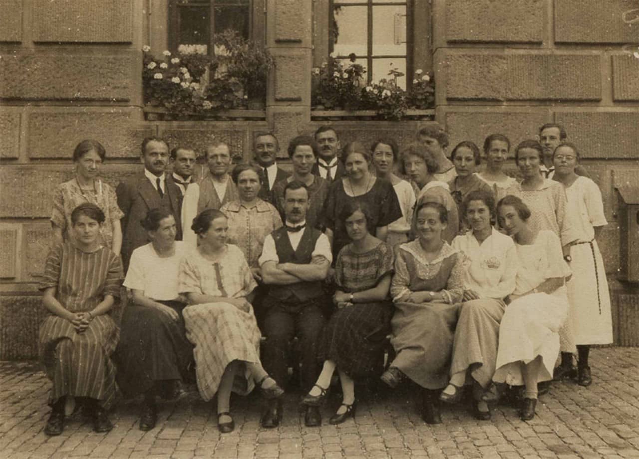 Schwarz-weiss Foto mit gelbstich einer Klasse am Lehrerinnenseminar in Aarau. In der ersten Reihe sitzen nur Frauen mit Ausnahme des Lehrers der in der Mitte sitzt. Zwei weitere Reihen von Männern und Frauen stehend hinten dran. Im Hintergrund ist eine Hausmauer mit zwei Fenstern zu sehen, welche Geranien auf dem Fenstersims haben. 