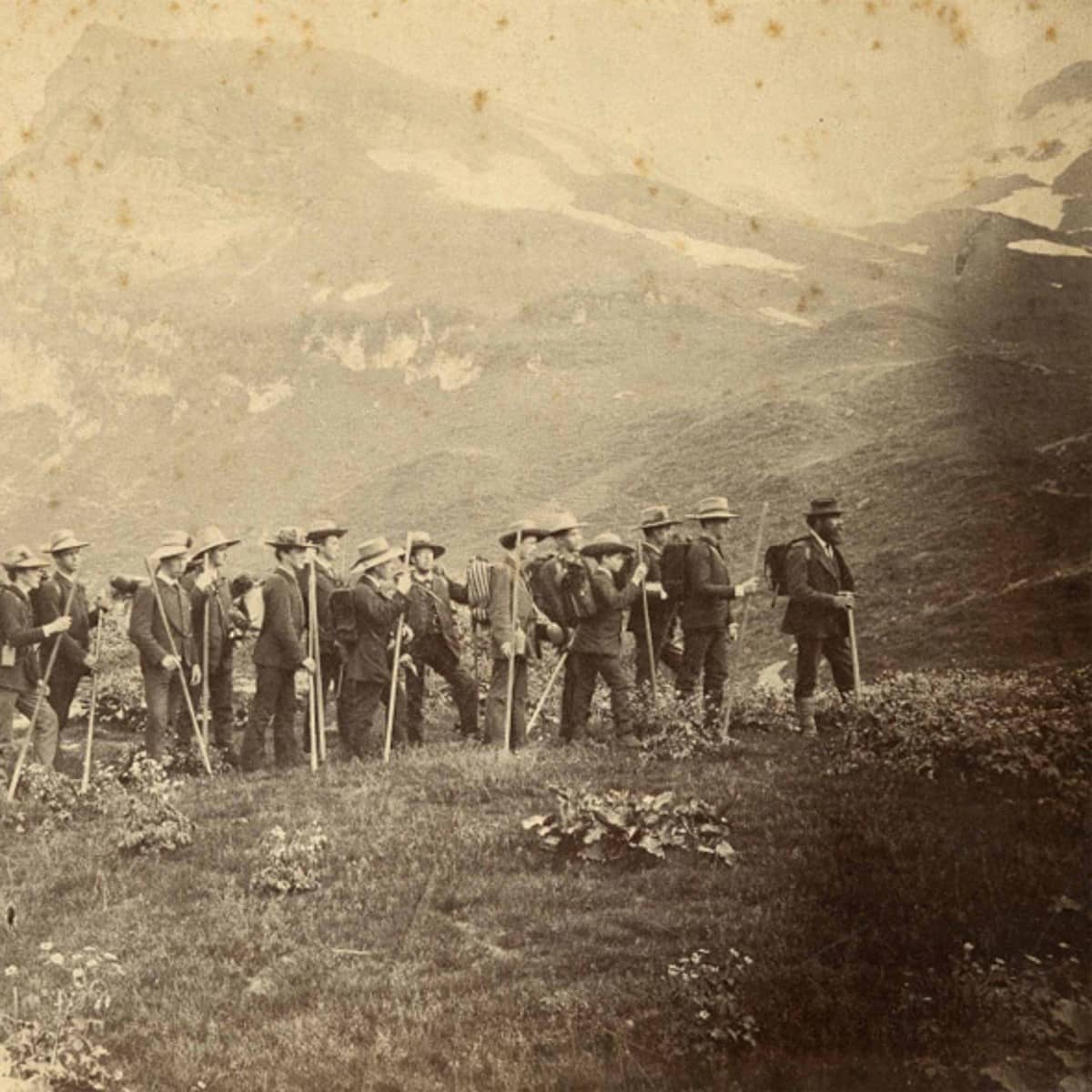 Schwarz-weiss Fotografie einer Gruppe von jungen Männern auf einer Wiese vor einem Bergrücken. Alle haben Wanderstöcke und Rucksäcke dabei und Hüte auf dem Kopf. 
