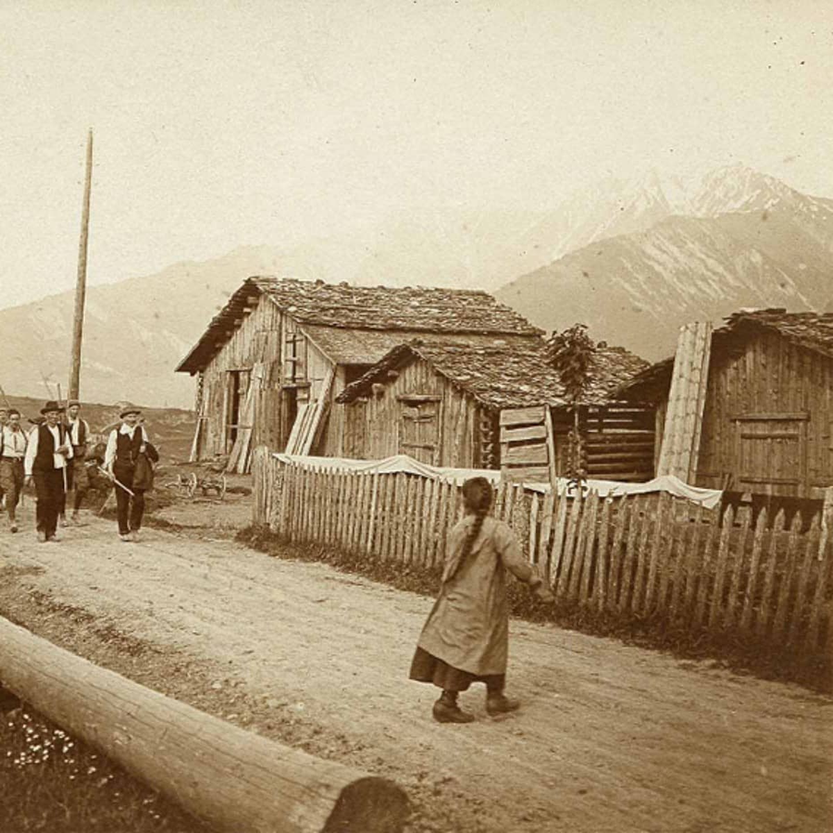 Schwarz-weiss Fotografie mit gelbstich. Links ist eine Gruppe von Personen mit Wanderstöcken im Anmarsch. Sie befinden sich auf einem Feldweg der an Holzhäusern und einem Holzzaun vorbeigeht. In der Bildmitte ist eine Frau mit langem Zopf von hinten zu sehen. Sie blickt der Gruppe entgegen und ihr Gesicht ist nicht erkennbar. 