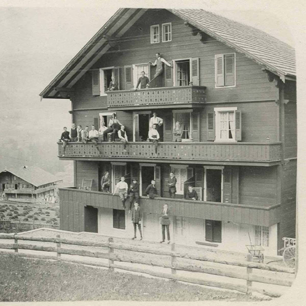 Schwarz-weiss Fotografie einer Aussenansicht eines grossen Holzhauses mit drei Stockwerken. Jeder Stock hat einen Balkon auf denen Schüler stehen und in die Kamera schauen. Im Hintergrund links ist ein weiteres Holzhaus zu sehen. 