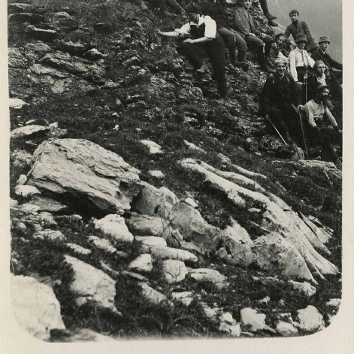 Schwarz-weiss Fotografie einer Wandergruppe die auf einem Bergabhang sitzt. Die Gruppe von Personen befindet sich am oberen rechten Rand des Bildes. Die Personen schauen entweder in die Kamera oder in die Ferne und von einigen sind nur die Beine erkennbar. Sie haben Hüte auf dem Kopf, Rucksäcke dabei und Wanderstöcke in der Hand. Im Vordergrund ist ein steiler Abhang mit Steinen und Felsen zu sehen. 