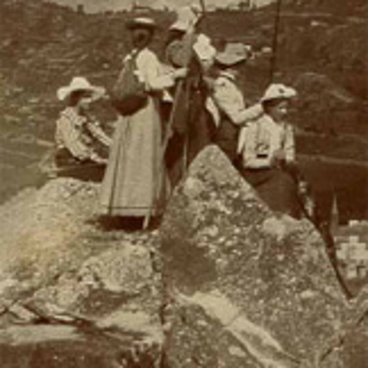 Schwarz-weiss Fotografie, leicht verschwommen, einer Gruppe von 5 Frauen die auf einem Fels stehen und sitzen. Die Frauen tragen lange Röcke, Hüte, Rucksäcke und haben Wanderstöcke in der Hand. Sie sind von hinten oder im Profil zu sehen und schauen nicht zum Betrachtenden. Im Hintergrund sind Häuser und eine Turmspitze sowie Berge zu sehen. Der Himmel ist wolkenverhangen. 