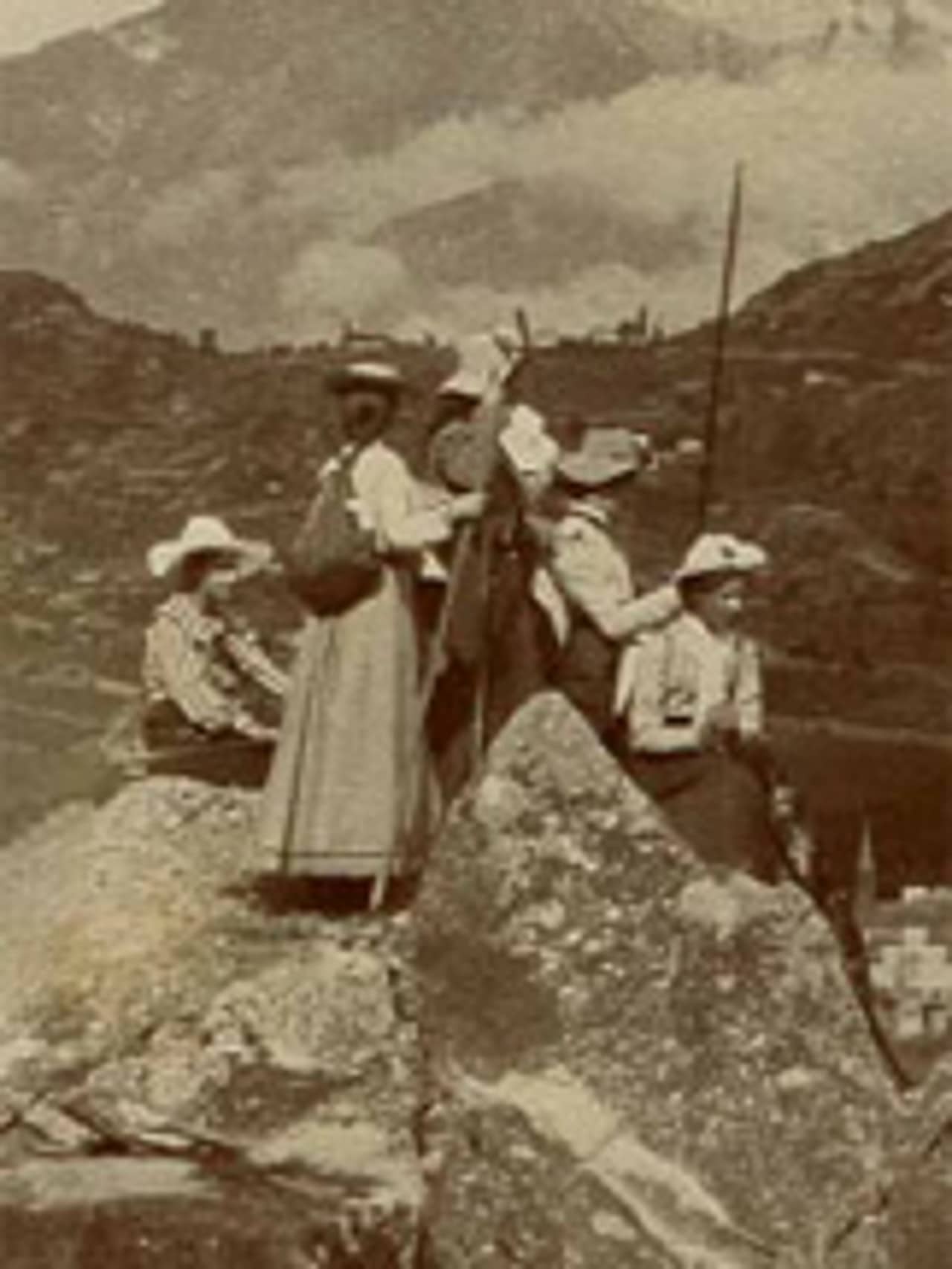 Schwarz-weiss Fotografie, leicht verschwommen, einer Gruppe von 5 Frauen die auf einem Fels stehen und sitzen. Die Frauen tragen lange Röcke, Hüte, Rucksäcke und haben Wanderstöcke in der Hand. Sie sind von hinten oder im Profil zu sehen und schauen nicht zum Betrachtenden. Im Hintergrund sind Häuser und eine Turmspitze sowie Berge zu sehen. Der Himmel ist wolkenverhangen.
