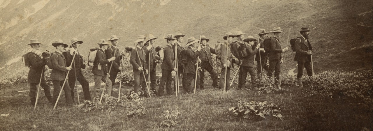 Schwarz-weiss Fotografie einer Gruppe von jungen Männern auf einer Wiese vor einem Bergrücken. Alle haben Wanderstöcke und Rucksäcke dabei und Hütte auf dem Kopf. 