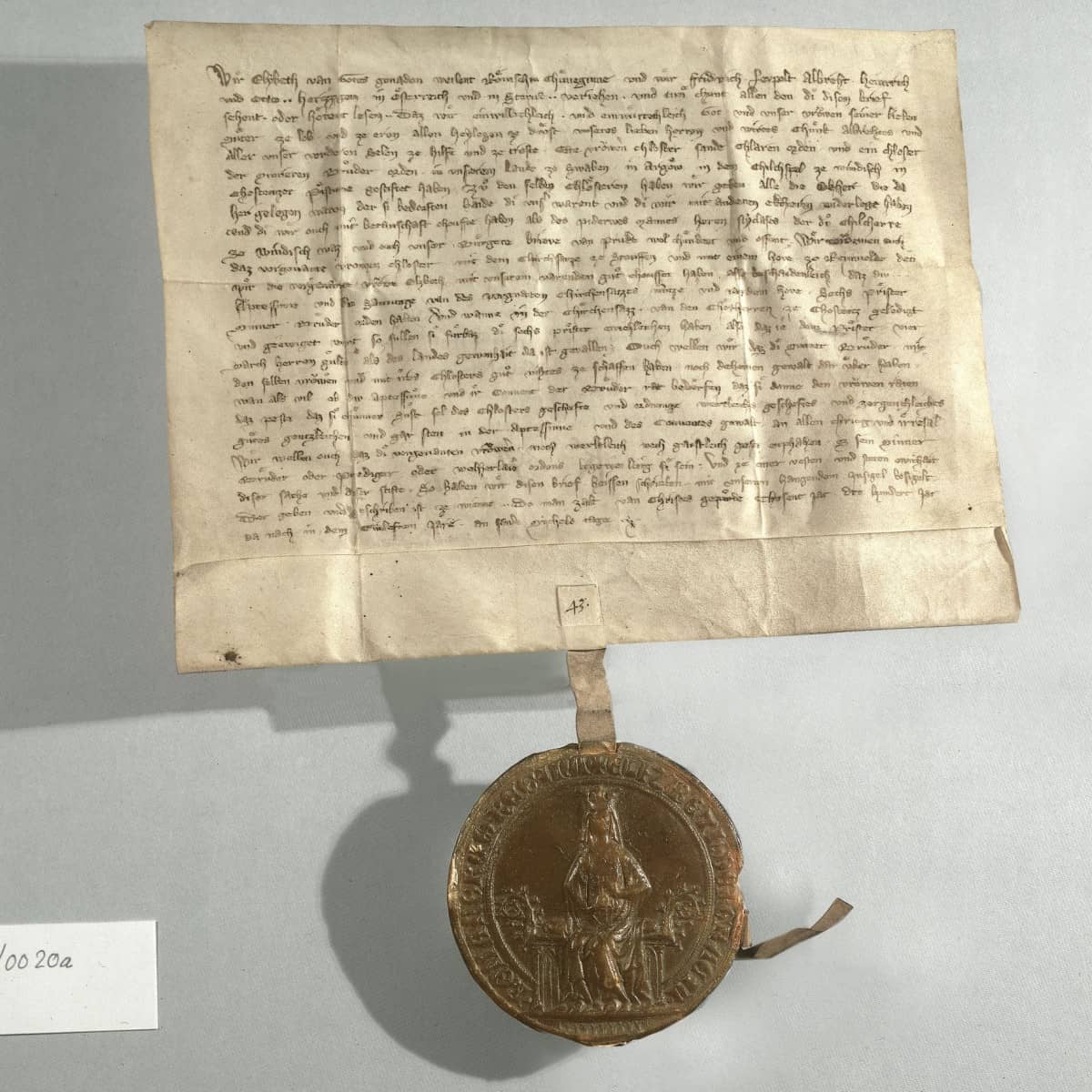 Pergament Urkunde mit dunkler Schrift und nach unten hängendem Siegel welches Königin auf Thron zeigt. Urkunde und Siegel werfen nach links einen Schatten.