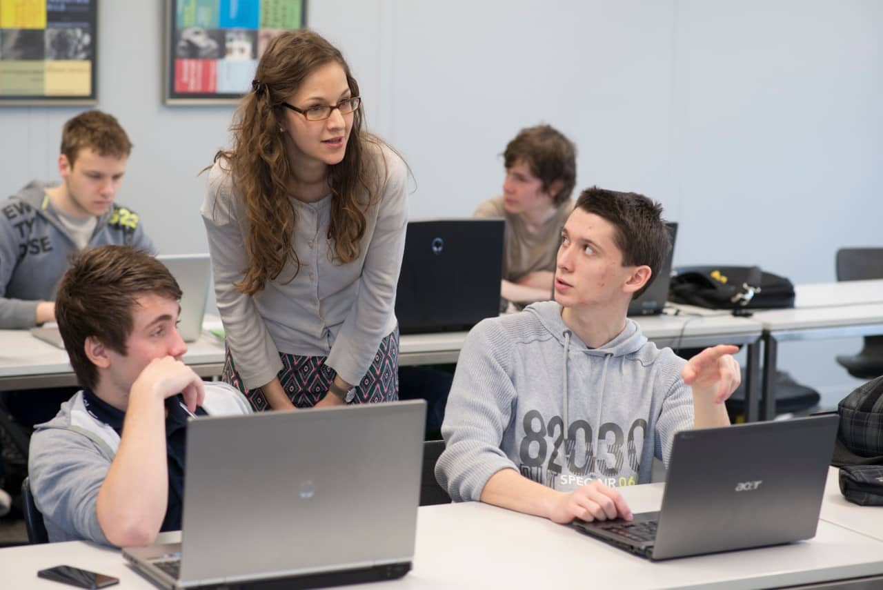 Zwei Schüler mit Laptops vor sich im Gespräch mit ihrer Lehrerin