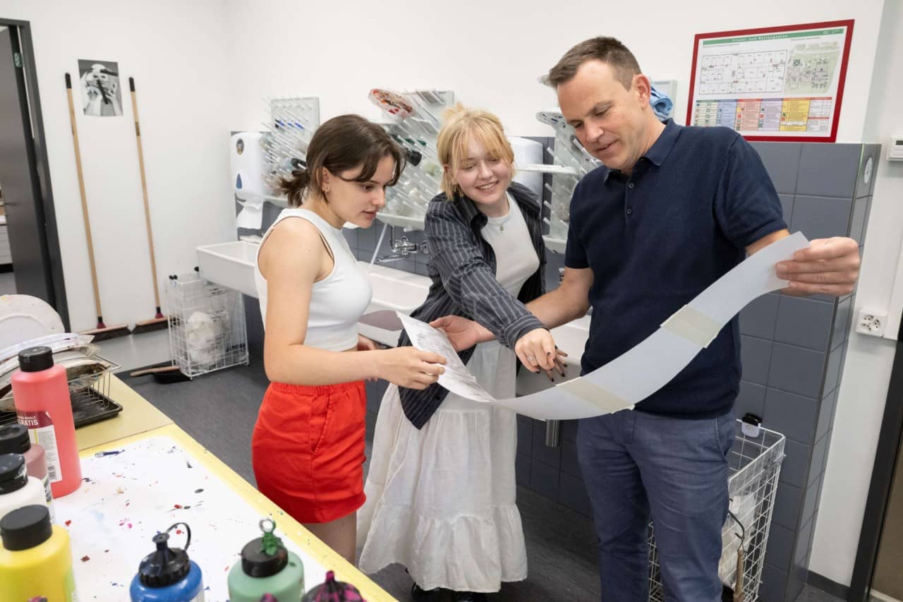 Zwei Schülerinnen und ein Mann halten Papierstreifen