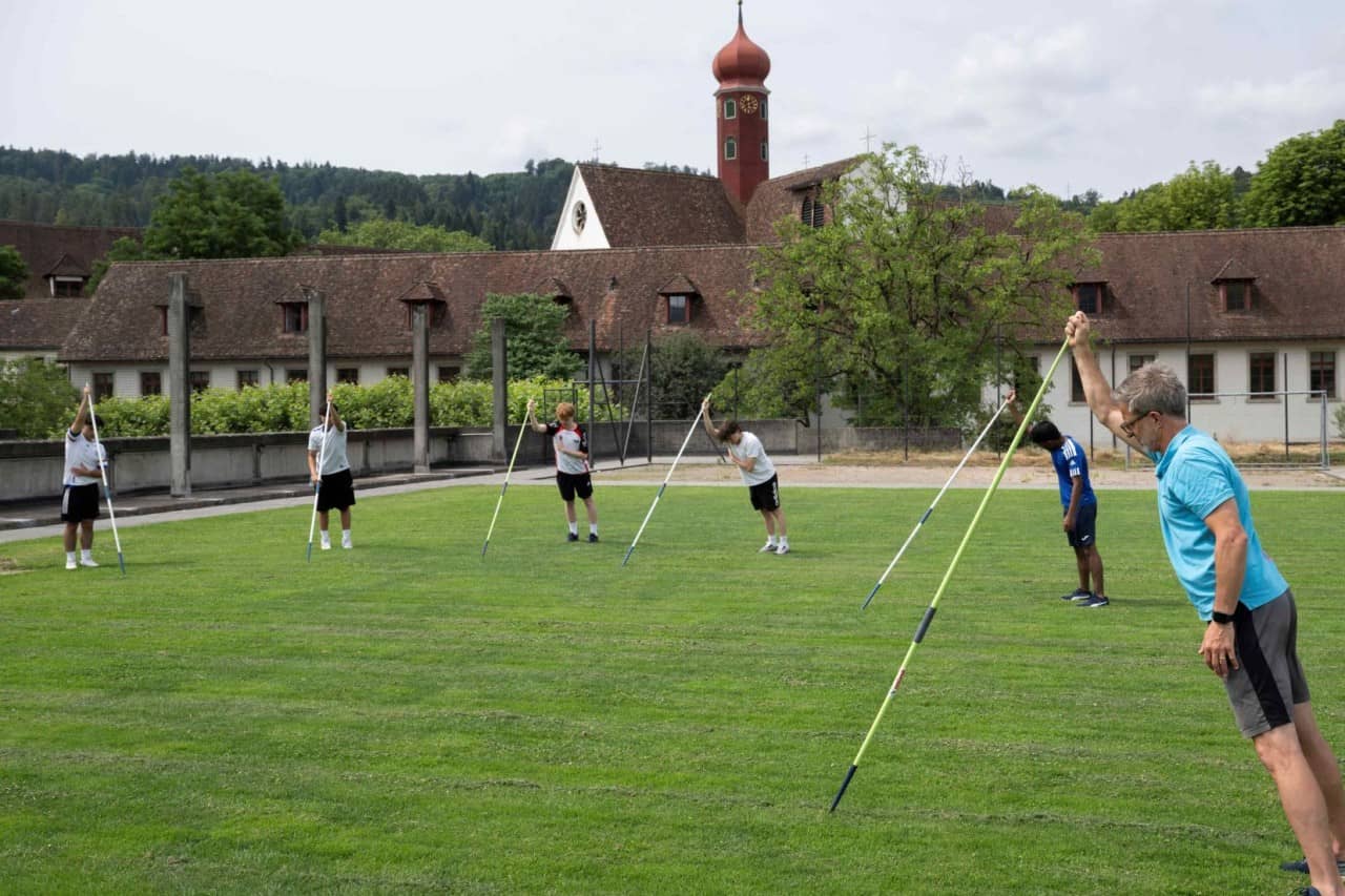 Sechs Personen halten Speere in Rasen gesteckt. Im Hintergrund sieht man Häuser.