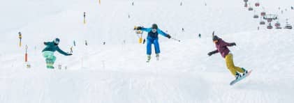 Ein Skifahrer/in und zwei Snowboarder/innen fahren auf der Piste den Hang hinunter.