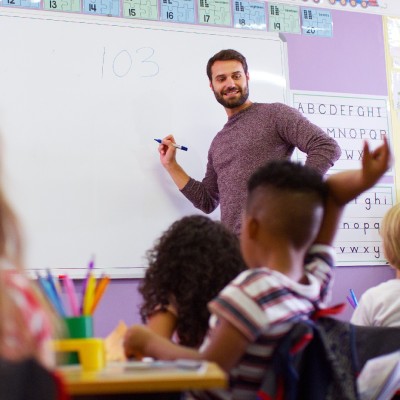 Lehrer steht mit Stift vor Wandtafel und schaut zur Klasse von der 4 Kinder zu sehen sind.
