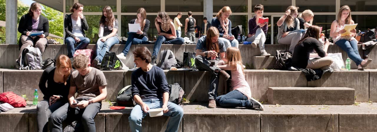 Jugendliche Schülerinnen und Schüler sitzen auf grossen Treppen vor einem Schulhaus und sind in Gesprächen oder in Hausaufgaben vertieft.