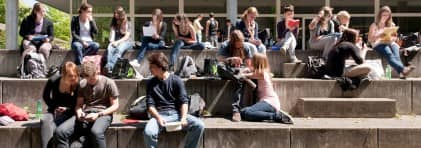Jugendliche Schülerinnen und Schüler sitzen auf grossen Treppen vor einem Schulhaus und sind in Gesprächen oder in Hausaufgaben vertieft.