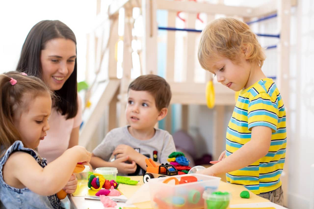 Eine Kindergartenlehrerin kniet neben drei Kindergartenkinder, die an einem Tisch spielen und basteln.