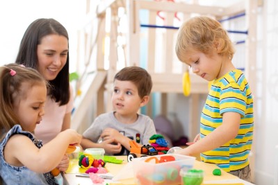 Eine Kindergartenlehrerin kniet neben drei Kindergartenkinder, die an einem Tisch spielen und basteln.