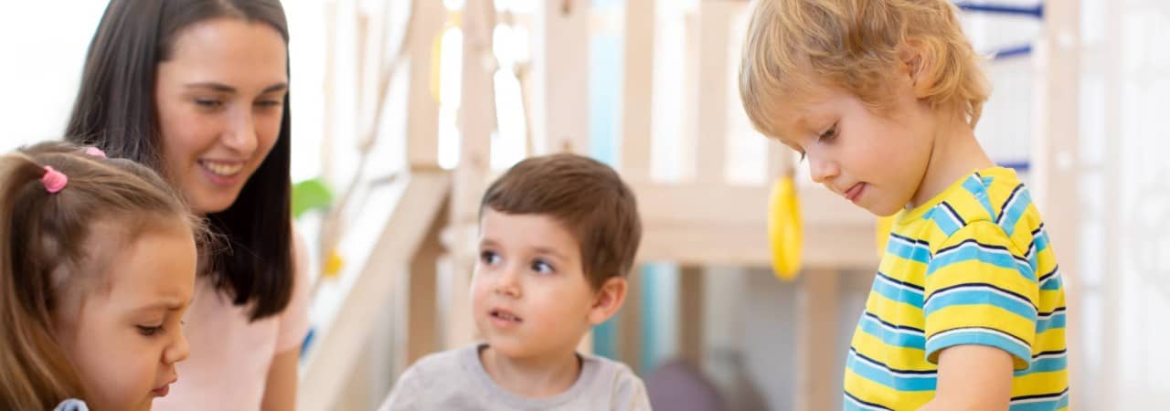 Eine Kindergartenlehrerin kniet neben drei Kindergartenkinder, die an einem Tisch spielen und basteln.