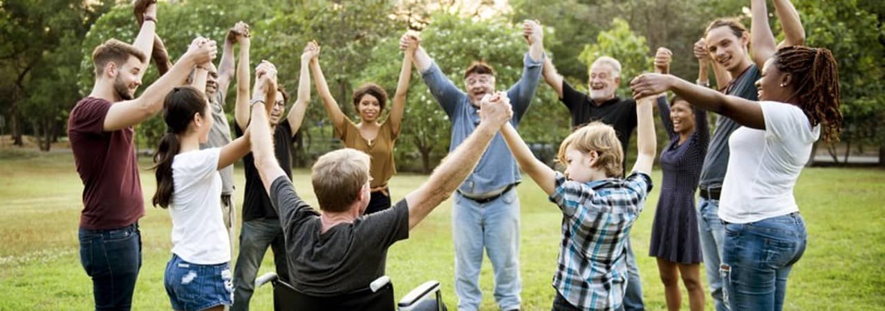 Eine Gruppe Menschen teils mit physischen Beeinträchtigung sind im Kreis auf einer grünen Wiese versammelt und halten sich mit erhobenen Armen an den Händen.