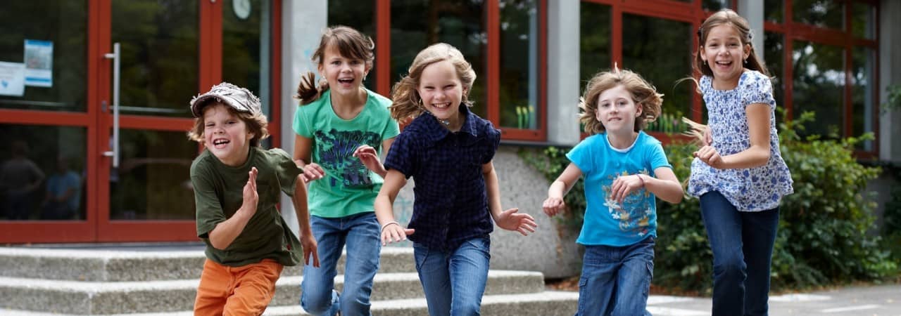 Fünf Schulkinder vor einem Schulgebäude rennen in Richtung des Fotografierenden.