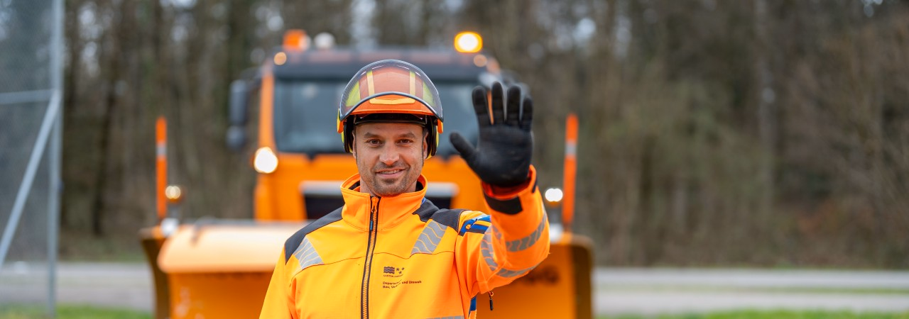 Männliche Person steht in Arbeitskleidung vor der linse und hält die linke Hand nach oben wie bei einem Halt-zeichen. Im Hintergrund ist ein oranger Schneepflug zu sehen. 