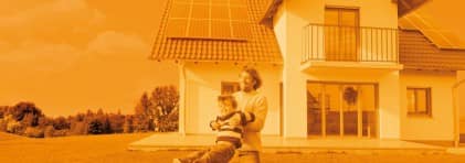 Im Hintergrund ein Einfamilienhaus mit Solarpanels auf dem Dach; im Vordergrund Spielt ein Vater mit seinem Sohn im Garten.