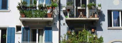 Fassade eines Mehrfamilienhauses mit Balkonen, auf denen verschiedene grüne und blühende Topfpflanzen stehen.