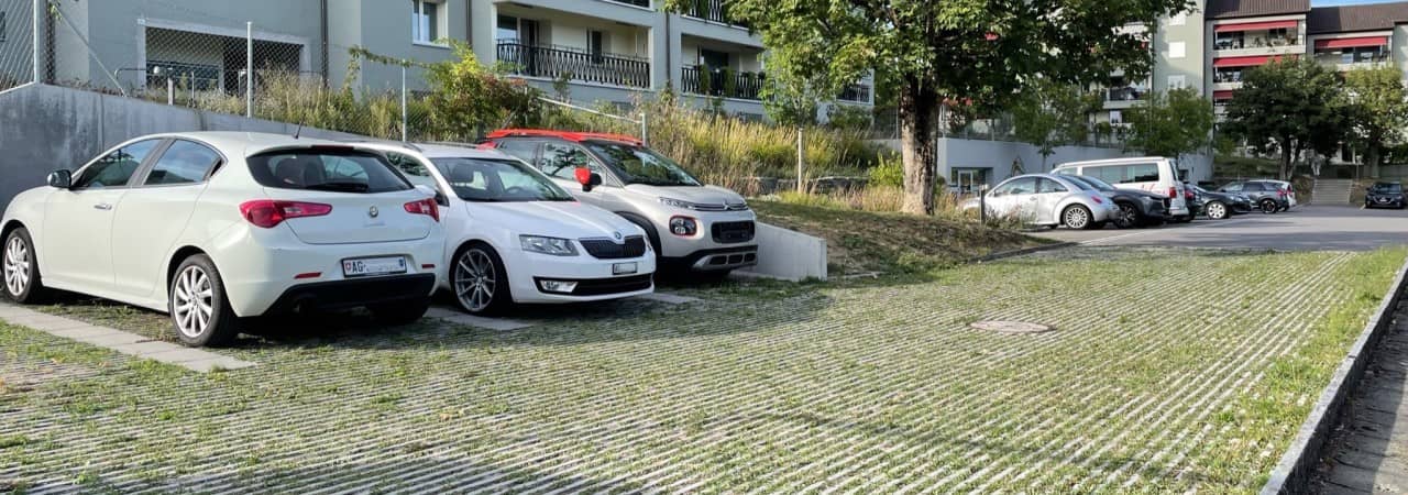 Entsiegelter Parkplatz mit parkierten Autos vor einem modernen Wohngebäude.