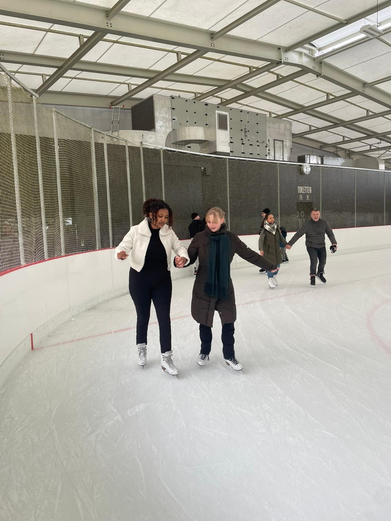 Mehrere Personen unterschiedlich jeden Alters beim Eislaufen auf einer Eisbahn; im Hintergrund eine graue Wand und Sanitärzeichen für Toiletten.