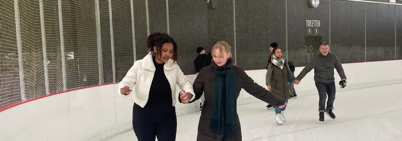 Mehrere Personen unterschiedlich jeden Alters beim Eislaufen auf einer Eisbahn; im Hintergrund eine graue Wand und Sanitärzeichen für Toiletten.