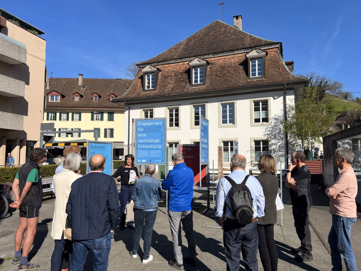 Dreizehn Personen stehen auf einem Platz und hören einer Rednerin zu; Infostele mit Text Engagement für eine nachhaltige Entwicklung vor historischem Gebäude.