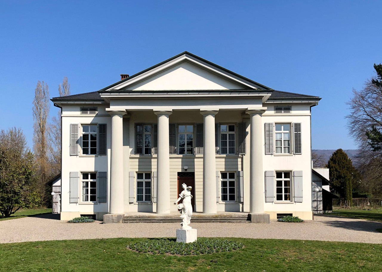 Frontansicht des Säulenhauses, im Vordergrund eine Statue einer Frau.