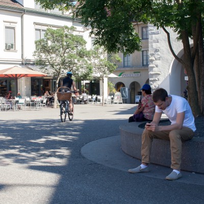 Zwei Personen sitzen im Schatten eines Baumes auf dem Schlossbergplatz in Baden, während eine Velofahrerin vorbeifährt.