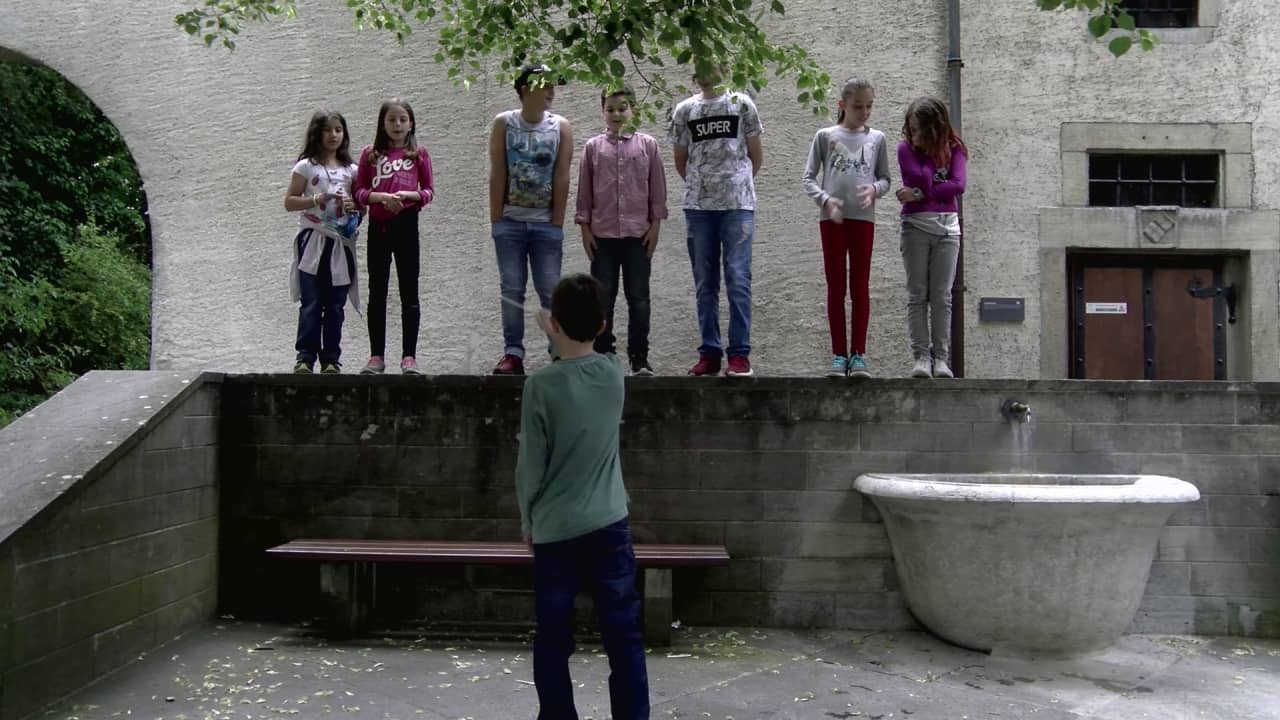 Kinder stehen singend auf einer Mauer, ein Kind vor ihnen dirigiert. 