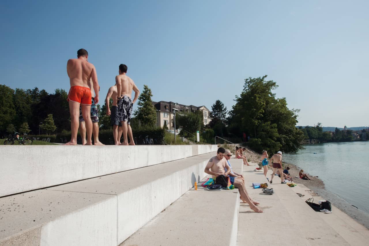 Menschen sitzen oder stehen auf in Badehosen auf breiten Stufen, welche zum Fluss führen